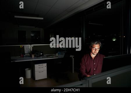 Middle aged businessman is sitting at a laptop in a dark office working on new deadline project - young male working late at night in the office Stock Photo