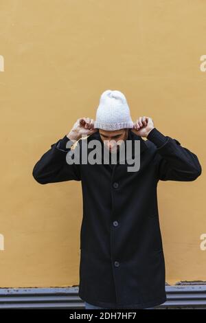 A vertical shot of a young man wearing a black coat and a white hat in front of a yellow wall Stock Photo