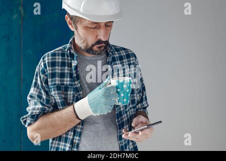 Construction engineer on a morning coffee break using mobile phone, relaxing and preparing for work Stock Photo