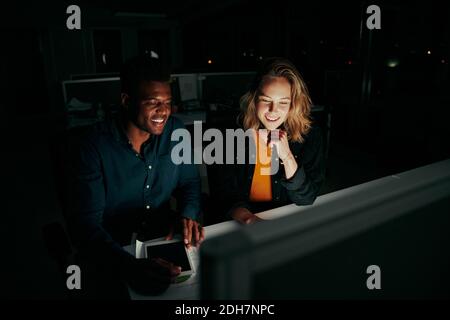 Two smiling young multiethnic business colleagues sitting together at desk working late at night in dark office on computer - two colleagues finishing Stock Photo
