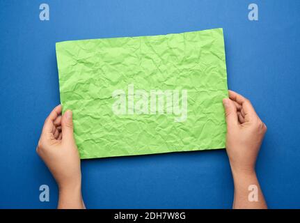 Two female hands are holding blank crumpled green sheet of paper on blue background Stock Photo