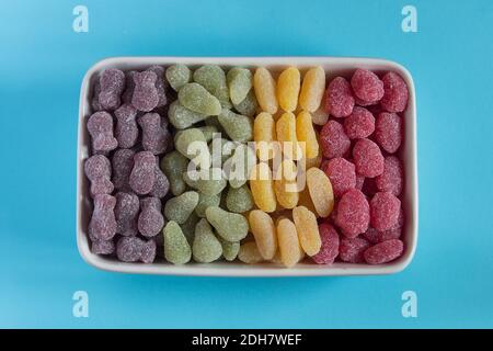 Rectangular ceramic bowl with sweets of different shapes ordered by color on a blue paper background. High-Sugar Foods. Stock Photo