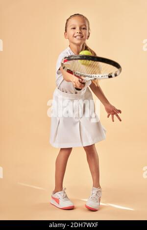 Cute female child in white tennis uniform bouncing tennis ball on racket. Isolated on light orange background Stock Photo