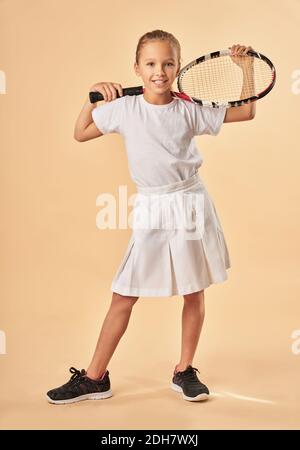 Adorable female child in tennis uniform looking at camera and smiling while holding tennis racket Stock Photo