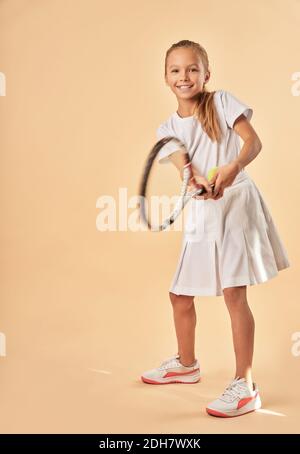 Adorable female child in tennis uniform looking at camera and smiling while holding racket Stock Photo