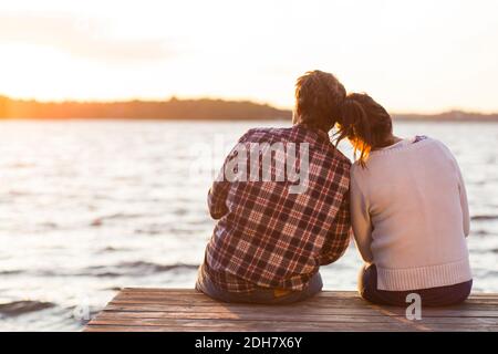 Rear view of loving couple sitting on pier against sea at sunset Stock Photo