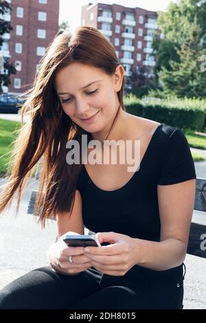 Smiling young woman using smart phone on bench outdoors Stock Photo