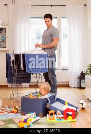 Father drying clothes while looking at baby boy playing with toys Stock Photo