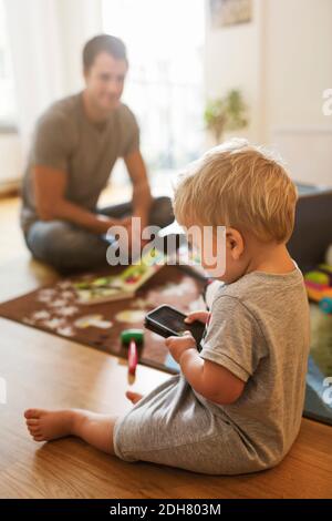 Baby boy using mobile phone on floor while father sitting with toys in background Stock Photo