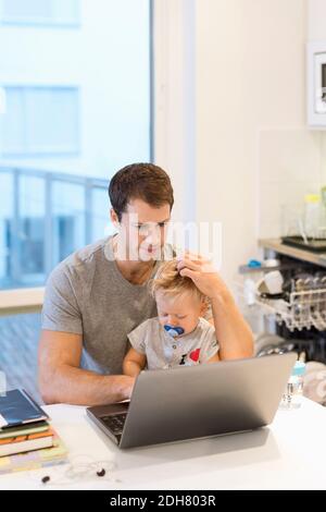 Baby boy sitting with father using laptop at table in house Stock Photo