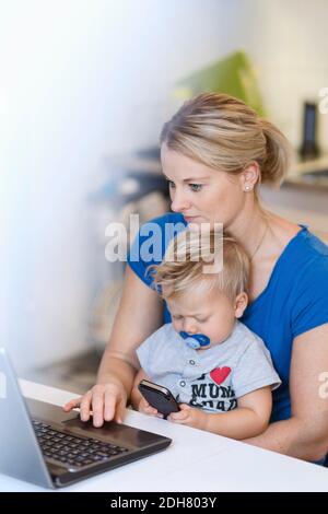 Mother working on laptop while baby using mobile phone at table Stock Photo