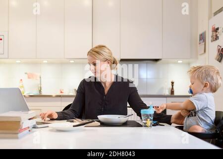 Mother working on laptop while taking care of baby at dining table Stock Photo