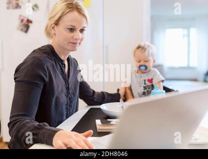 Woman working on laptop while taking care of baby boy at dining table Stock Photo