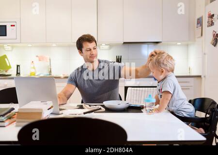 Father stroking baby boy while using laptop at dining table Stock Photo