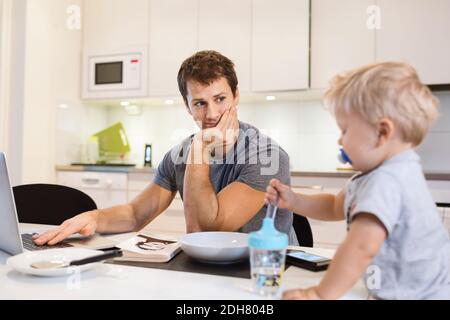 Father looking at baby boy while using laptop in kitchen Stock Photo