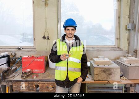 Portrait of confident young manual worker holding machine part in factory Stock Photo