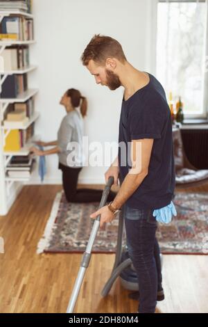 Side view of man vacuuming floor while woman cleaning shelves in background at home Stock Photo