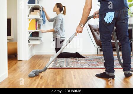Low section of man vacuuming floor while woman cleaning shelves in background at home Stock Photo