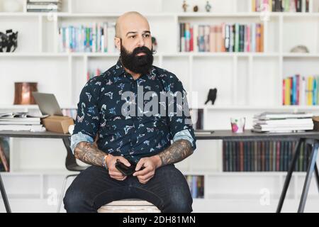Thoughtful male architect holding mobile phone while sitting in home office Stock Photo