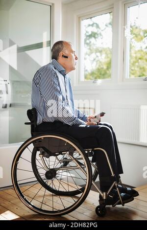 Mature man holding mobile phone while using Bluetooth in wheelchair at home Stock Photo