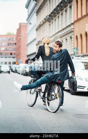 Rear view portrait of happy businessman sitting on back seat while female colleague riding bicycle Stock Photo