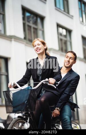 Happy businesswoman riding bicycle while colleague sitting on back seat outdoors Stock Photo