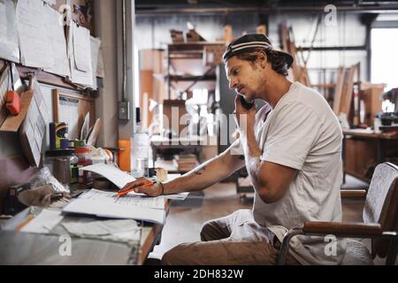 Side view of carpenter using mobile phone while reading document in workshop Stock Photo
