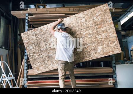 Rear view of man carrying wooden plank in front of shelf at workshop Stock Photo