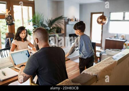 Rear view of man working on laptop while family at dining table in house Stock Photo