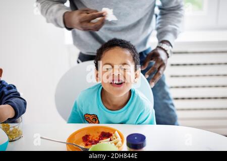 Boy making face while having food at table with father standing in background Stock Photo