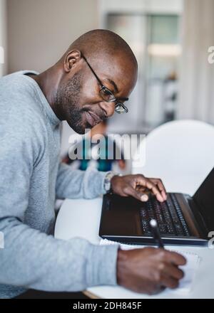 Side view of man writing while working on laptop at home Stock Photo