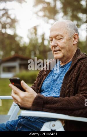 Senior gay man using smart phone sitting on chair at yard Stock Photo ...