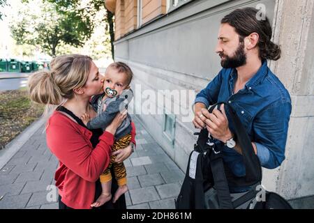 Working mother kissing baby boy while man holding carrier on sidewalk Stock Photo