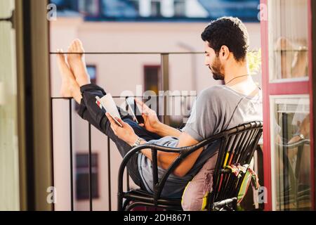 Side view of man using smart phone while holding guidebook at balcony Stock Photo