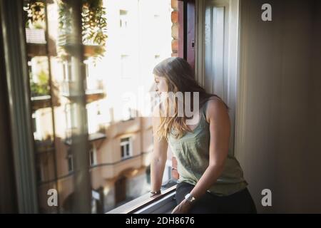 Young woman looking through window at home Stock Photo