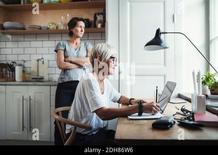 Industrial designer using laptop at table with colleague standing in kitchen Stock Photo