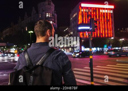 Rear view of man carrying backpack while standing on city street at night Stock Photo