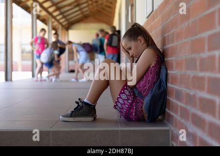 Schoolgirl sitting on the ground against a wall alone in the schoolyard at elementary school Stock Photo