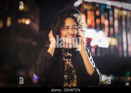 Happy young woman looking away while answering smart phone in city at night Stock Photo