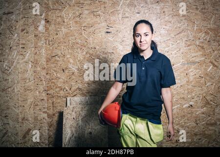 Portrait of confident female carpentry student holding hardhat while standing against wooden wall Stock Photo