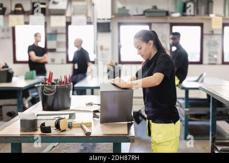 Side view of female student working on sheet metal with coworkers in background Stock Photo