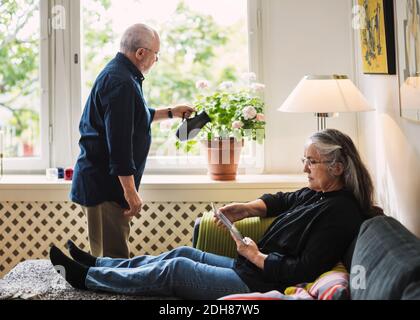 Full length of senior woman using digital tablet while man watering flower plant at home Stock Photo