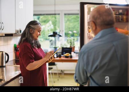 Side view of senior woman using digital tablet while man standing at refrigerator Stock Photo
