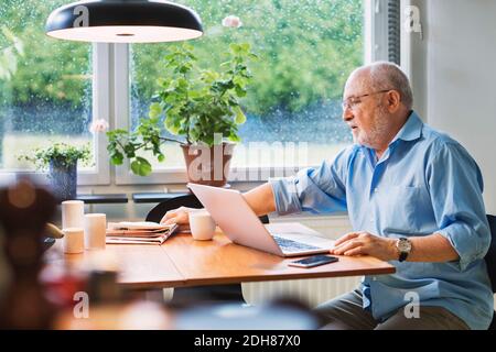 Senior man with laptop reaching for newspaper at table Stock Photo
