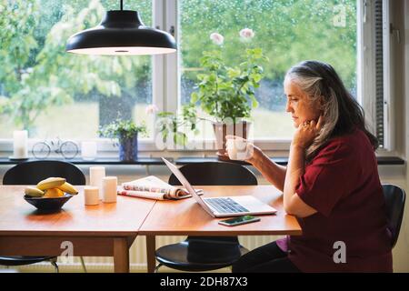 Side view of senior woman using laptop while having coffee at table Stock Photo