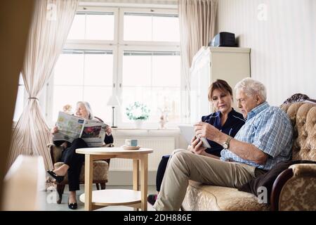 Senior man and caretaker using digital tablet while woman reading newspaper in nursing home Stock Photo