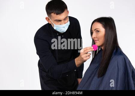A male professional makeup artist working with beautiful young woman wearing medical mask, with a cosmetic sponge. Stock Photo