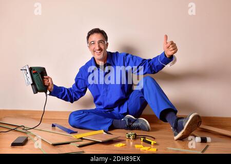 Professional parquet assembler with ok gesture and cutting machine in hand sitting on the floor assembling parquet. Horizontal composition. Stock Photo