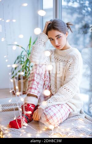 A young brunette woman dressed in a warm white sweater sits near the window and expects a miracle. Bokeh. Stock Photo