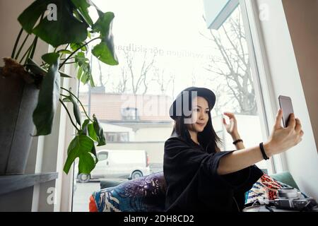 Smiling young woman taking selfie while sitting against window in office Stock Photo
